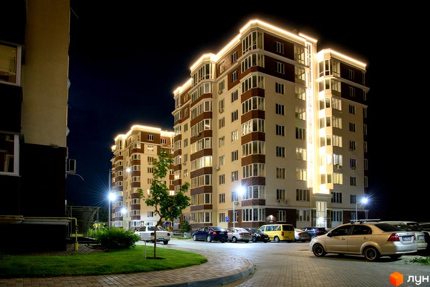 Житловий комплекс Золота Ера вночі з підсвічуванням. На території парковка з автомобілями та зелені насадження.