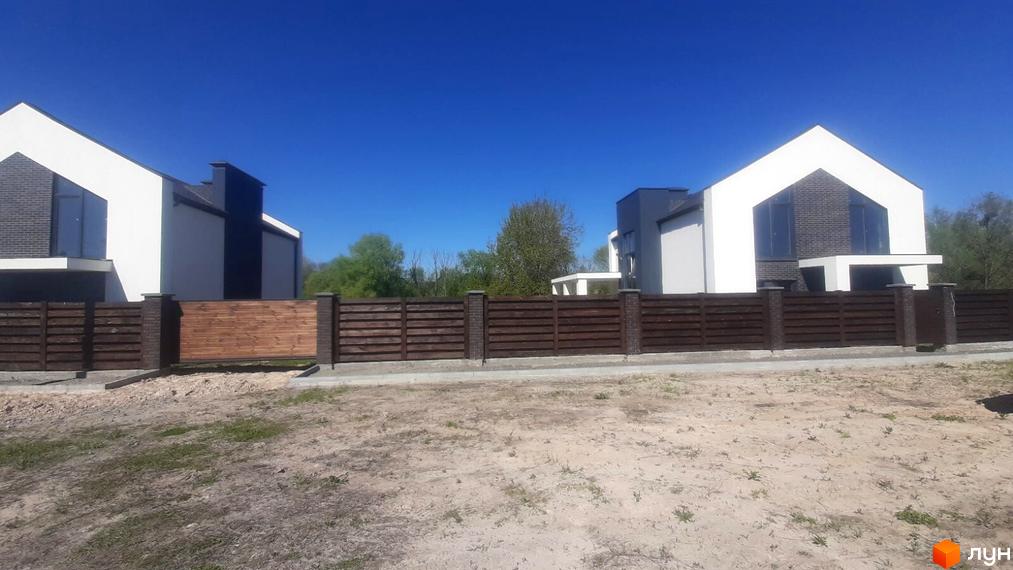 Два котеджі Котеджі біля Озер з білими фасадами та темною цеглою, огороджені коричневим парканом на піщаній ділянці.