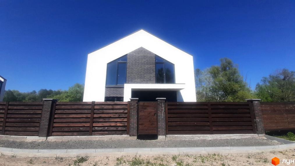 Білий будинок з великими вікнами та фасадом з темно-сірої цегли. Дерев'яний паркан та зелені насадження навколо будинку.