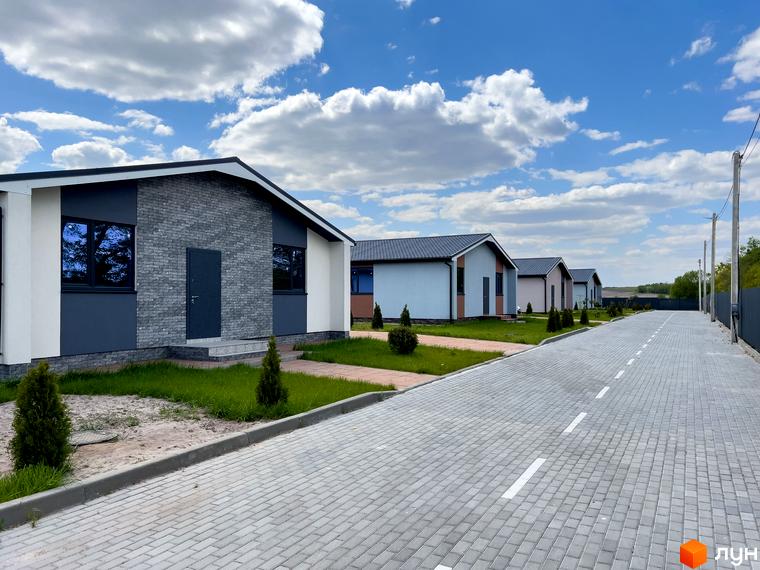 Одноповерхові будинки з сірими фасадами та темними дахами розташовані вздовж вимощеної дороги. Навколо будинків зелені газони та невеликі дерева.