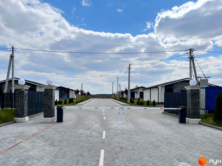 В'їзд до Котеджного містечка Millennium House з шлагбаумом та доріжкою з бруківки. По обидва боки розташовані одноповерхові будинки з темними дахами та комбінованими фасадами.