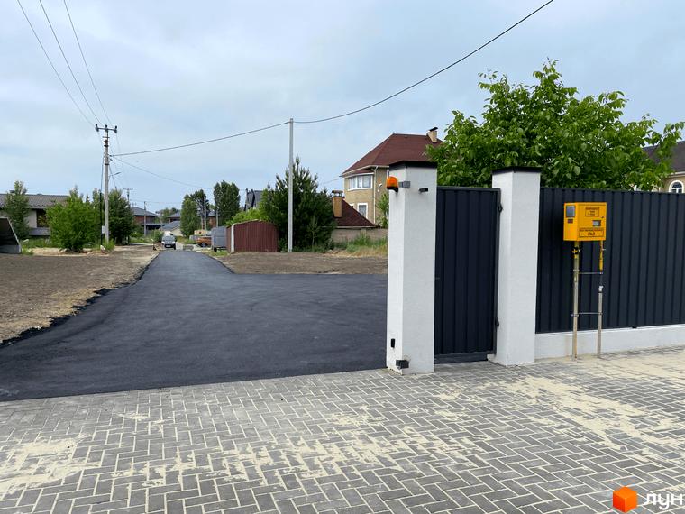 В'їзд до житлового комплексу з асфальтованою дорогою та котеджами на задньому плані. Огороджена територія з воротами та газовим лічильником.