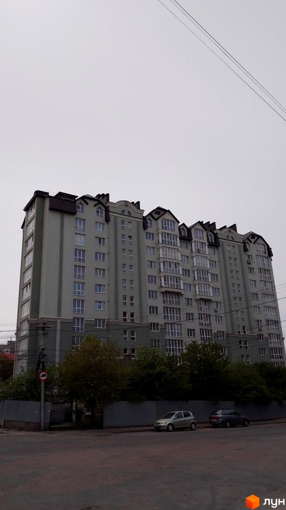 Висотна будівля Житлового комплексу з великою кількістю вікон та балконів. Перед будинком дерева та припарковані автомобілі.