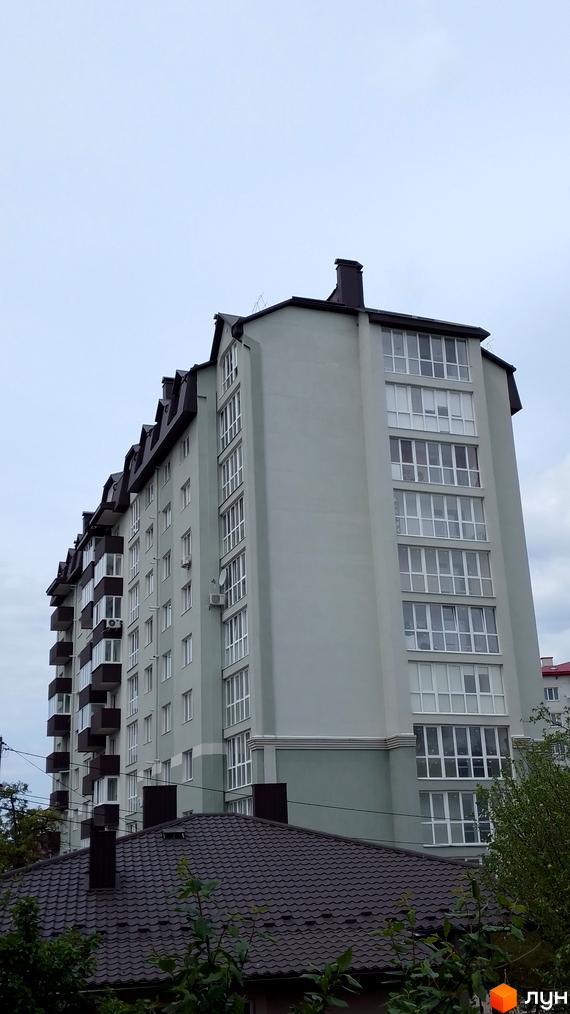 Висотна будівля Житловий комплекс з панорамними вікнами та балконами. На передньому плані видно дах будинку та зелені насадження.