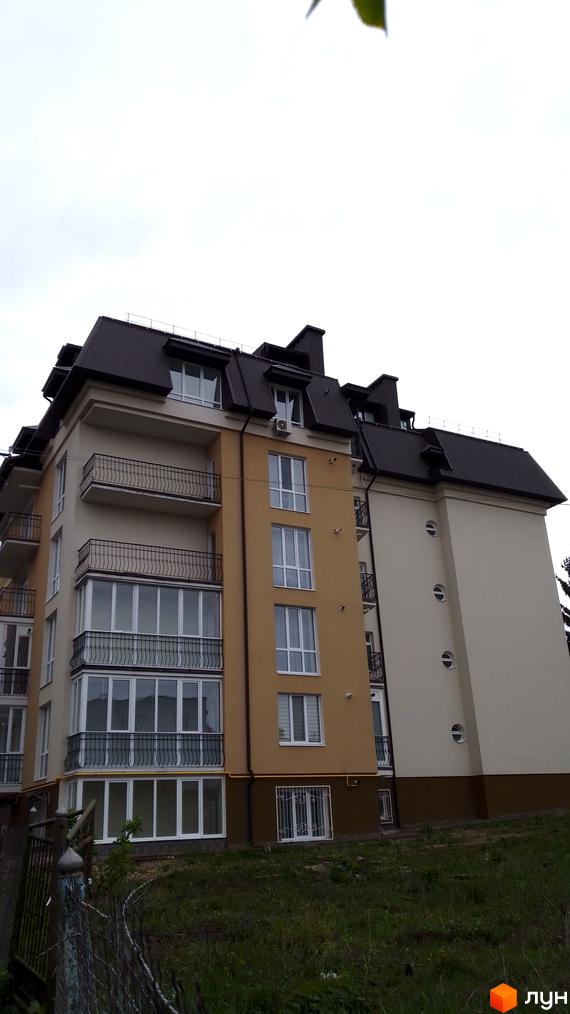 Багатоповерховий будинок з балконами та панорамними вікнами. На передньому плані видно огорожу та зелену прибудинкову територію.