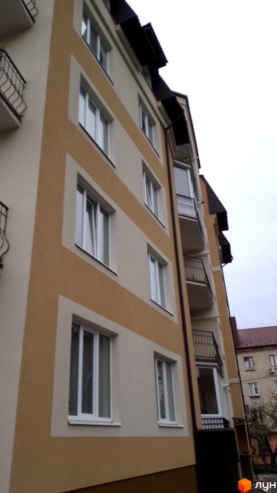 Фасад багатоповерхового будинку світло-бежевого кольору з коричневими декоративними смугами, білі вікна та балкони з чорними кованими перилами.