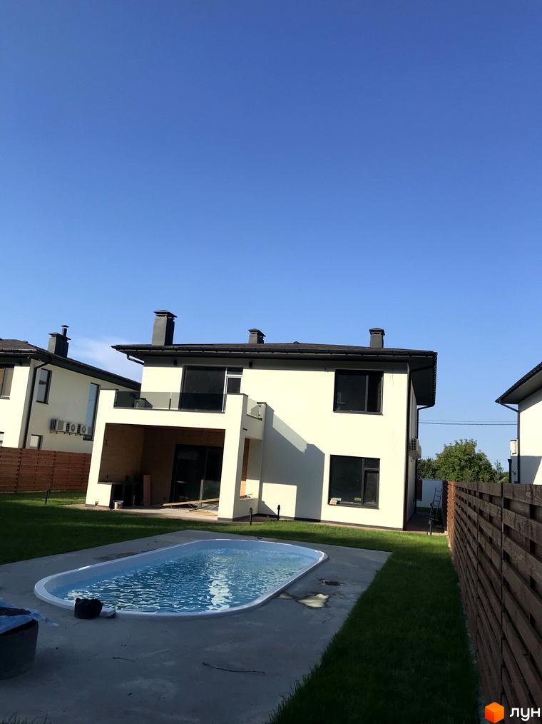 Зовнішній вигляд світлих двоповерхових будинків з темними дахами та вікнами. Поруч розташований басейн та зелена галявина. Котеджне містечко Lugovoy.