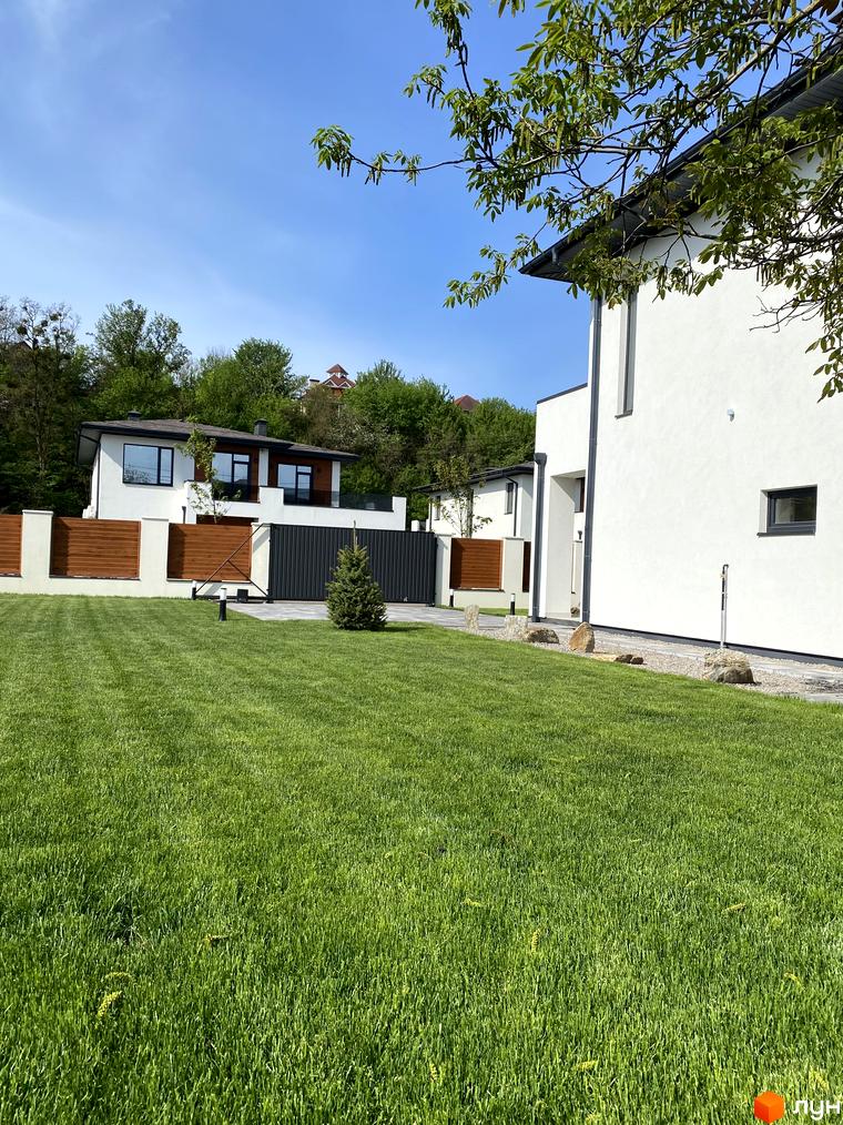 Котеджне містечко Lugovoy: білий двоповерховий будинок з великими вікнами та доглянутою зеленою галявиною перед ним. Територія огороджена дерев'яним парканом та має сучасні ворота.