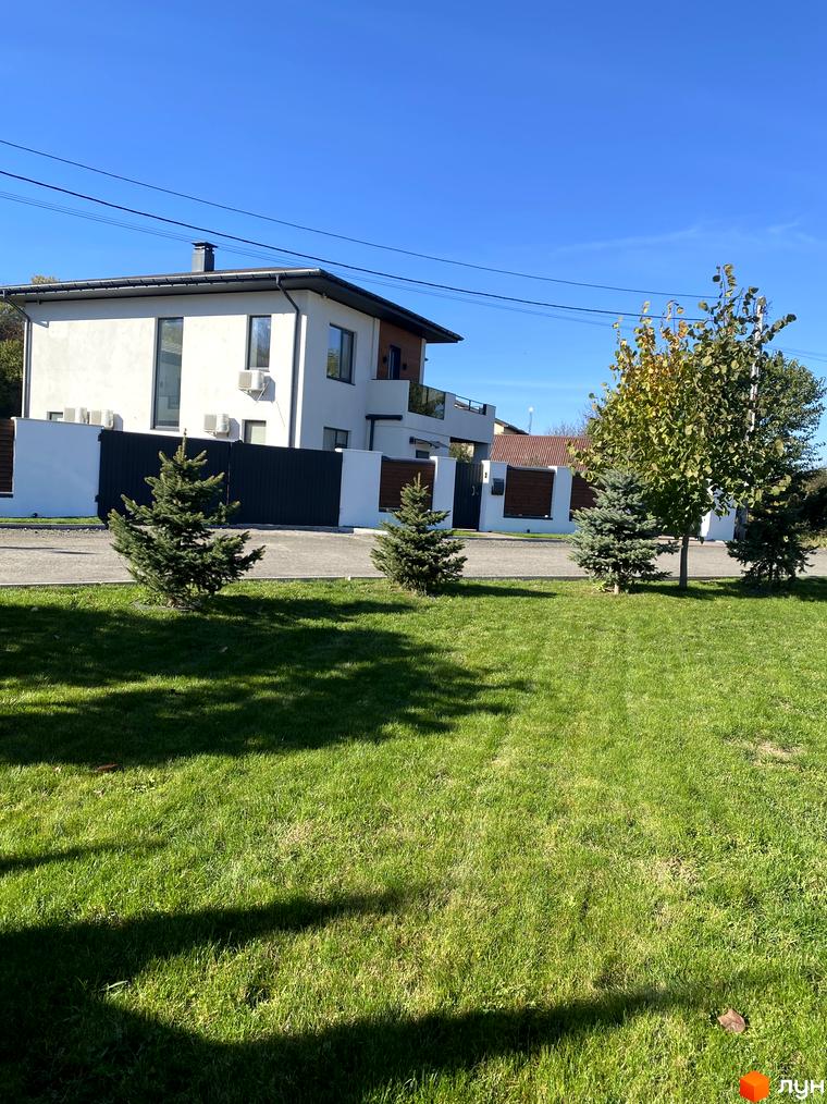 Котеджне містечко Lugovoy. Зовнішній вигляд білого двоповерхового будинку з темним дахом та дерев'яними елементами. Прибудинкова територія з зеленим газоном, ялинками та деревами.