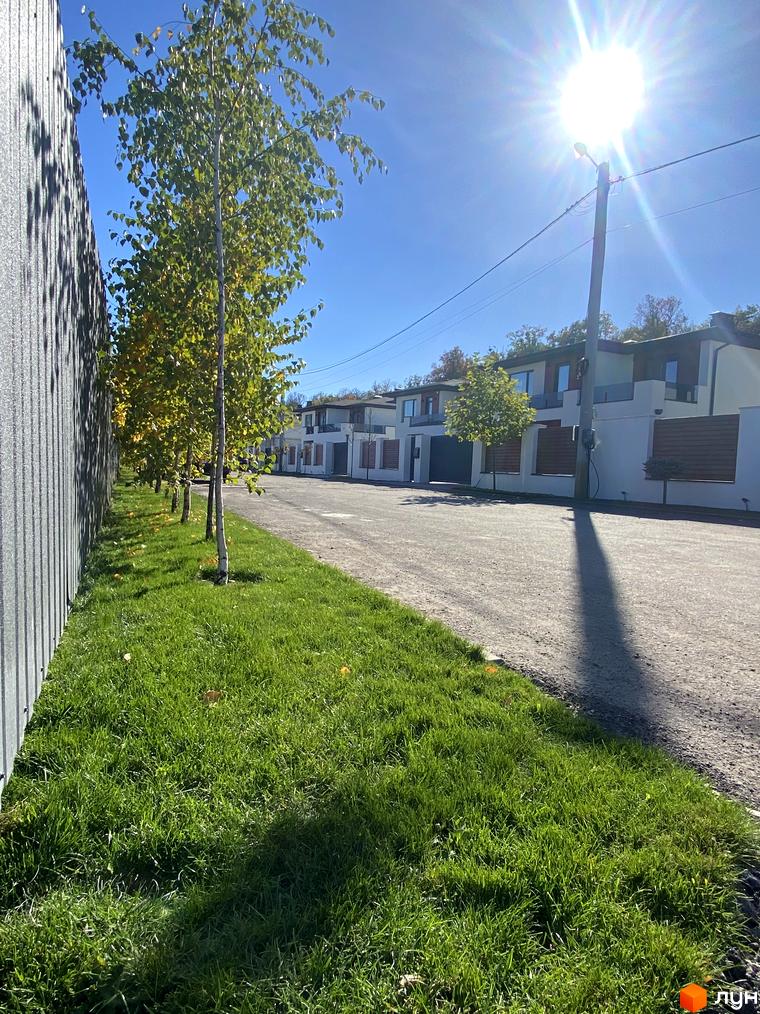 Котеджне містечко Lugovoy: фасади білих будинків з темними вікнами та дерев'яними елементами. Доглянута зелена галявина з молодими деревами вздовж дороги.