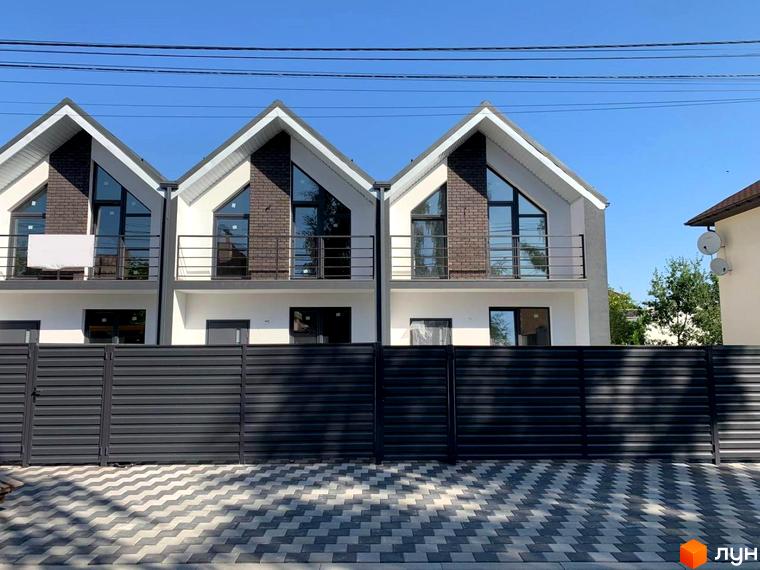 Три таунхауси з білими фасадами, панорамними вікнами та чорними балконами. Перед будинками розташований паркан та тротуар з бруківки.