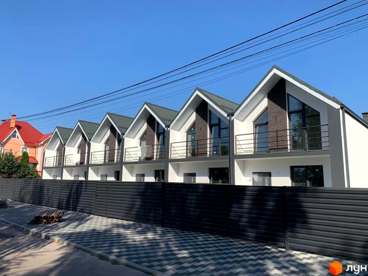 Зображення сторінки житлового комплексу Таунхаус Vasylkivska. Білі будинки з сірими дахами, балконами та чорним парканом, розташовані вздовж тротуару з плиткою.