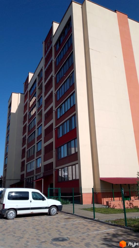 Висотна будівля Житлового комплексу з великими вікнами та світлим фасадом. На передньому плані припаркований автомобіль, територія огороджена.
