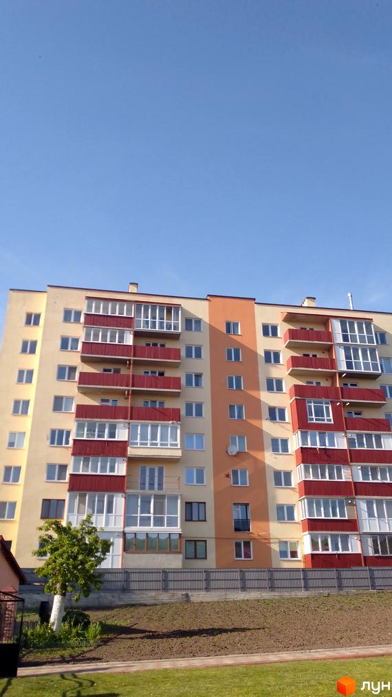 Багатоповерховий житловий будинок з жовто-помаранчевим фасадом та червоними балконами. Перед ним розташовані огорожа, ділянка землі та зелена трава.