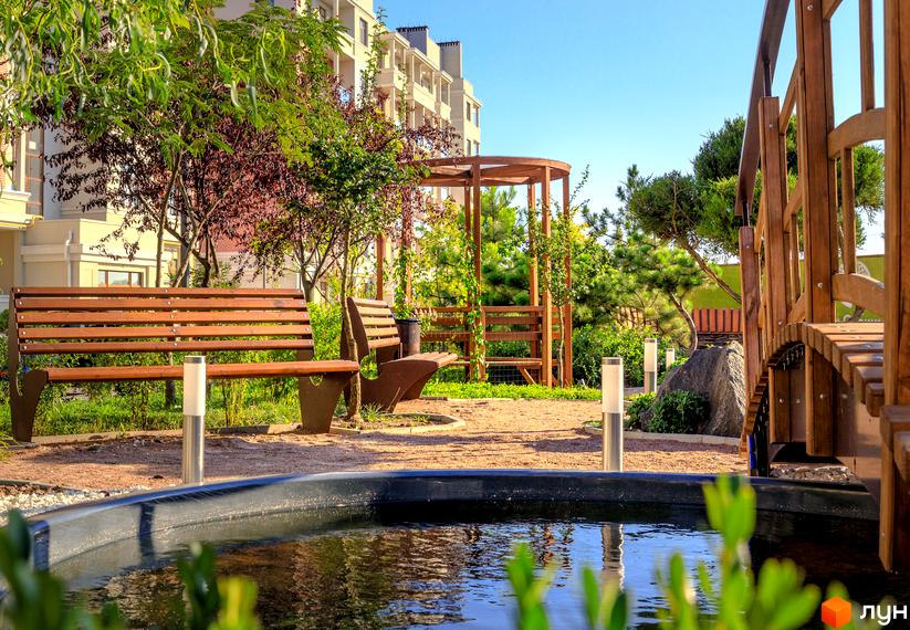 Житловий комплекс Парк Совіньйон. Затишна прибудинкова територія з ландшафтним дизайном, альтанкою, містком та водоймою. Навколо зелені насадження та сучасна будівля.