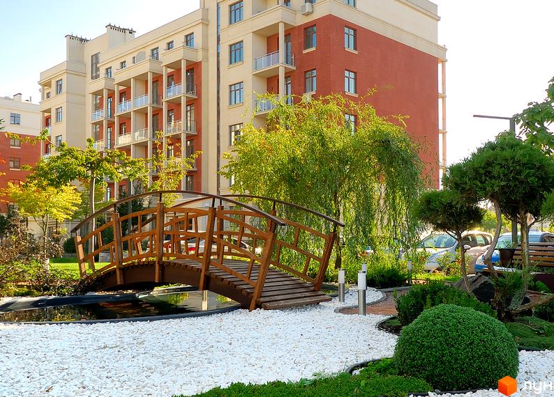 Житловий комплекс Парк Совіньйон. Фасад будинку з червоної цегли та бежевих елементів, балкони зі скляними огорожами. Прибудинкова територія з доглянутим ландшафтом, містком через водойму та декоративними рослинами.