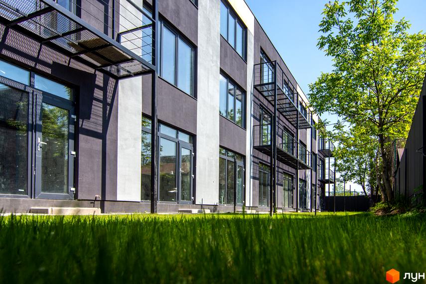 Фасад Таунхауси Rusaniv Gardens з темними стінами, великими вікнами та металевими балконами. Перед будинком зелений газон і дерева.