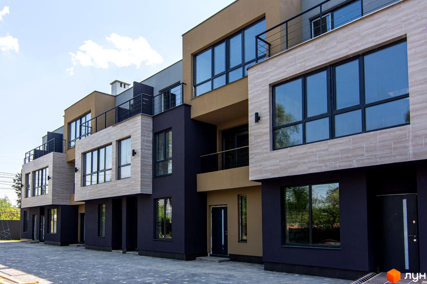 Ряд таунхаусів Rusaniv Gardens з фасадами темно-сірого, бежевого та світлого кольорів, великими вікнами та балконами. Прибудинкова територія викладена сірою бруківкою.