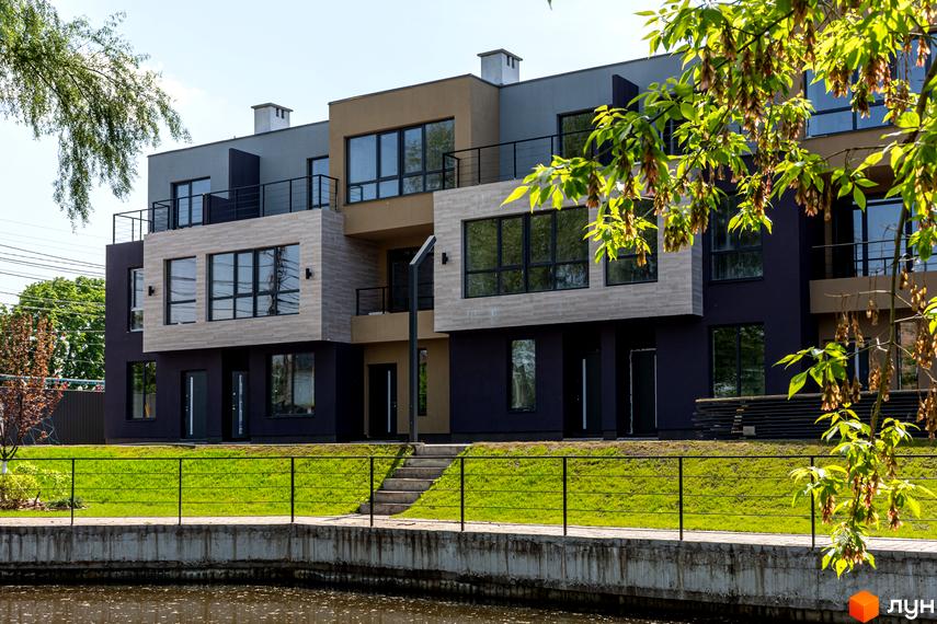 Таунхауси Rusaniv Gardens з багатотонними фасадами, великими вікнами та балконами. Перед ними зелений газон, огорожа та водойма.