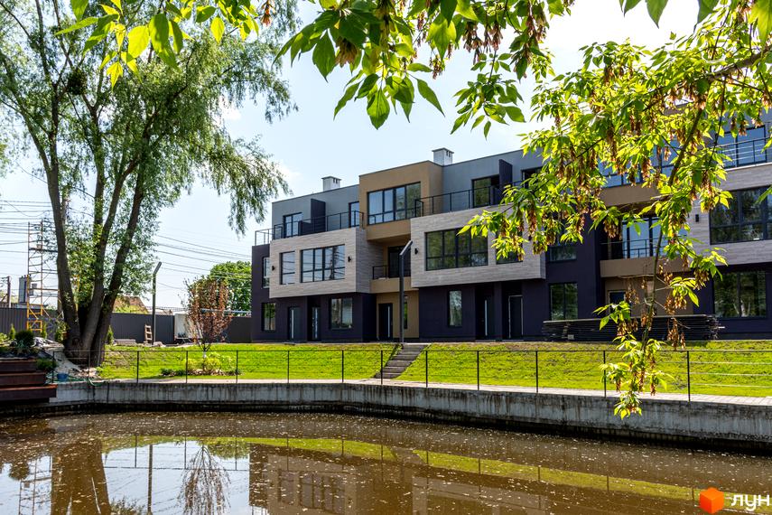 Триповерховий будинок з великими вікнами та балконами на березі водойми. Поруч газон з огорожею та дерева.