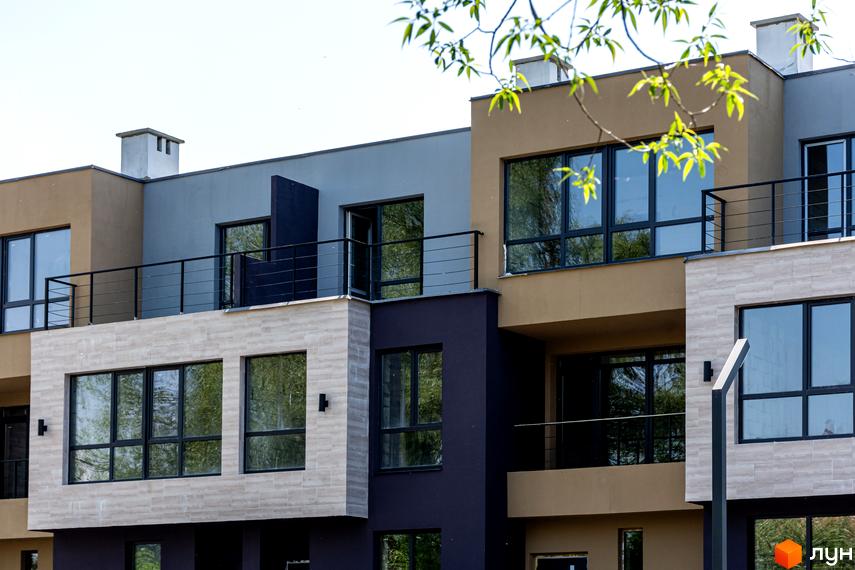 Зовнішній вигляд Таунхаусів Rusaniv Gardens з багатосекційним фасадом у бежевих, сірих та темно-фіолетових тонах, великими вікнами та балконами. На передньому плані видно зелені гілки дерев.