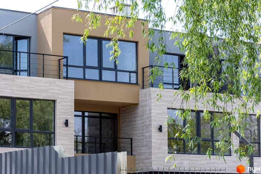 Зображення сторінки житлового комплексу Rusaniv Gardens. Двоповерховий будинок з великими вікнами та балконом, облицьований світлою плиткою. На передньому плані паркан та зелень.