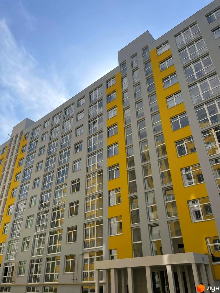 Висотна будівля житлового комплексу з жовтими та сірими фасадами. На передньому плані видно вхідну групу з колонами.