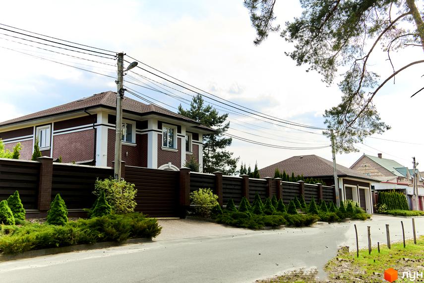 Будинок з коричневою черепицею та цегляною кладкою, огороджений парканом. На прибудинковій території висаджені кущі та дерева, асфальтована дорога.