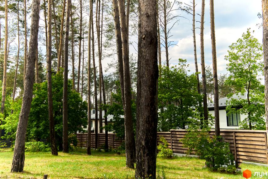 Зображення будинку в Містечку котеджів Квітка Полонини. Навколо будинку зелена трава та дерева, за будинком видно паркан.