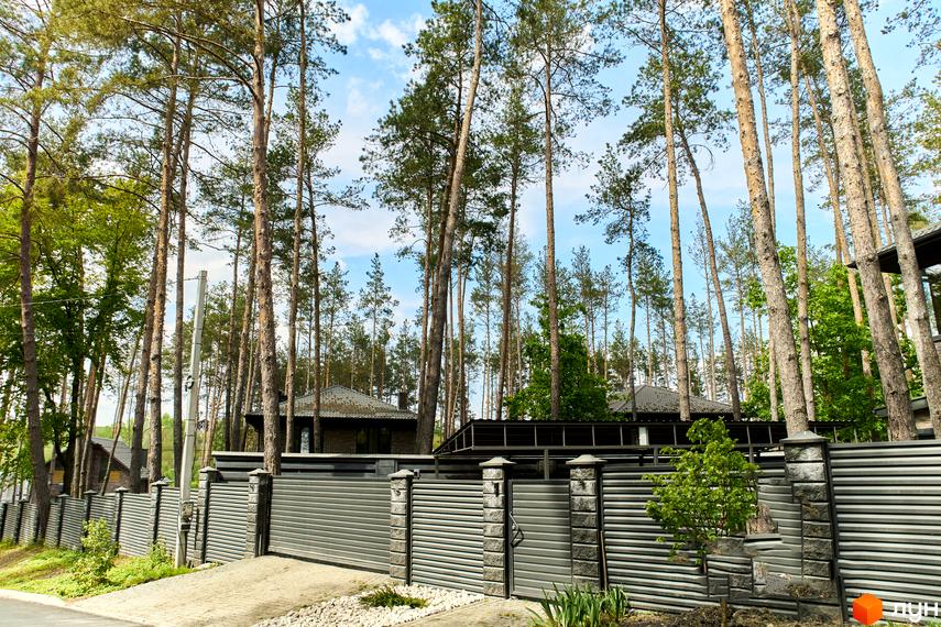 Вид на будинки Котеджне містечко Квітка Полонини, за високим сірим парканом та деревами. На території є зелені насадження та бруківка.