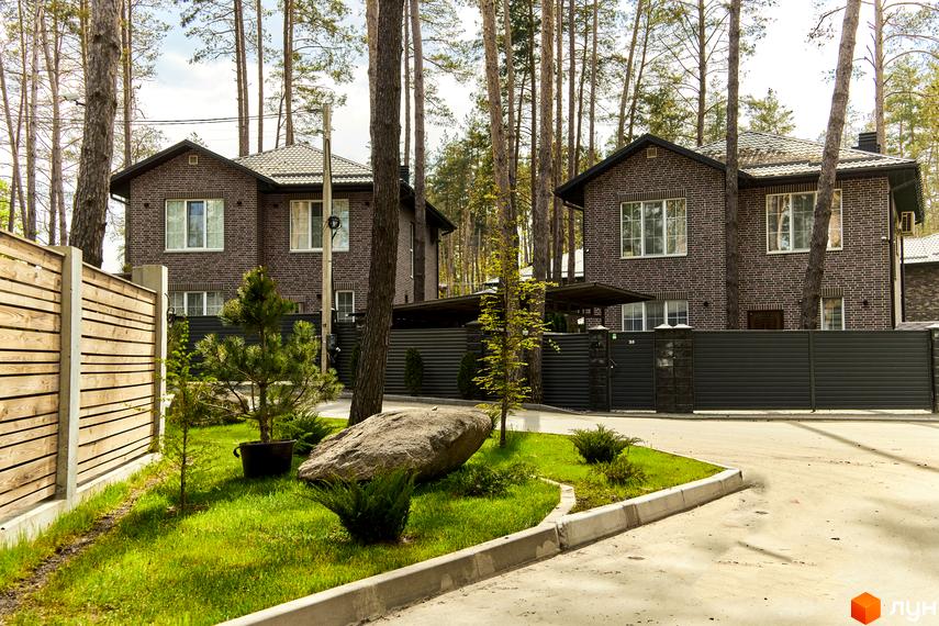 Двоповерховий будинок з цегляним фасадом, великими вікнами та темним дахом. На території Котеджне містечко Квітка Полонини є зелені насадження та бруківка.