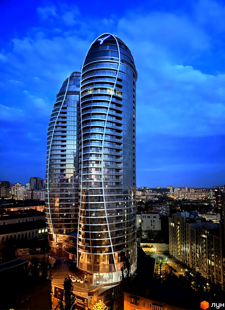 Житловий комплекс Taryan Towers з футуристичною архітектурою та неоновим підсвічуванням на тлі нічного неба. Навколо будівлі розташоване міське оточення з іншими новобудовами.