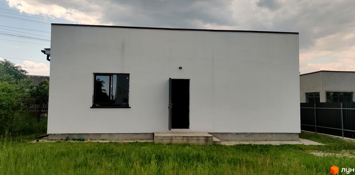 Білий одноповерховий будинок Котеджного містечка Любимівка з темним дахом, вікном та відкритими дверима. Перед будинком зелена трава.