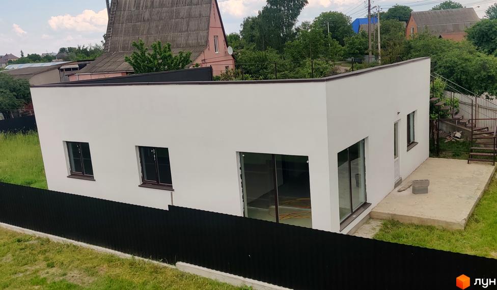 Котеджне містечко Любимівка: Білий одноповерховий будинок з темними вікнами. Прилегла територія з травою, парканом та бетонним майданчиком.