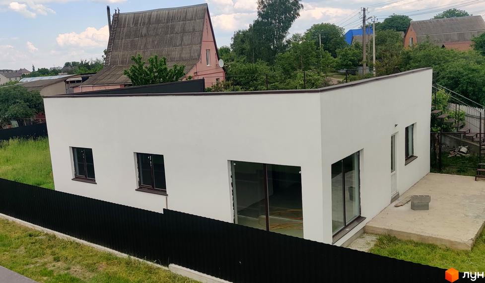 Котеджне містечко Любимівка: Білий одноповерховий будинок з темними вікнами та великими скляними дверима. Поруч бетонна тераса та чорний паркан.