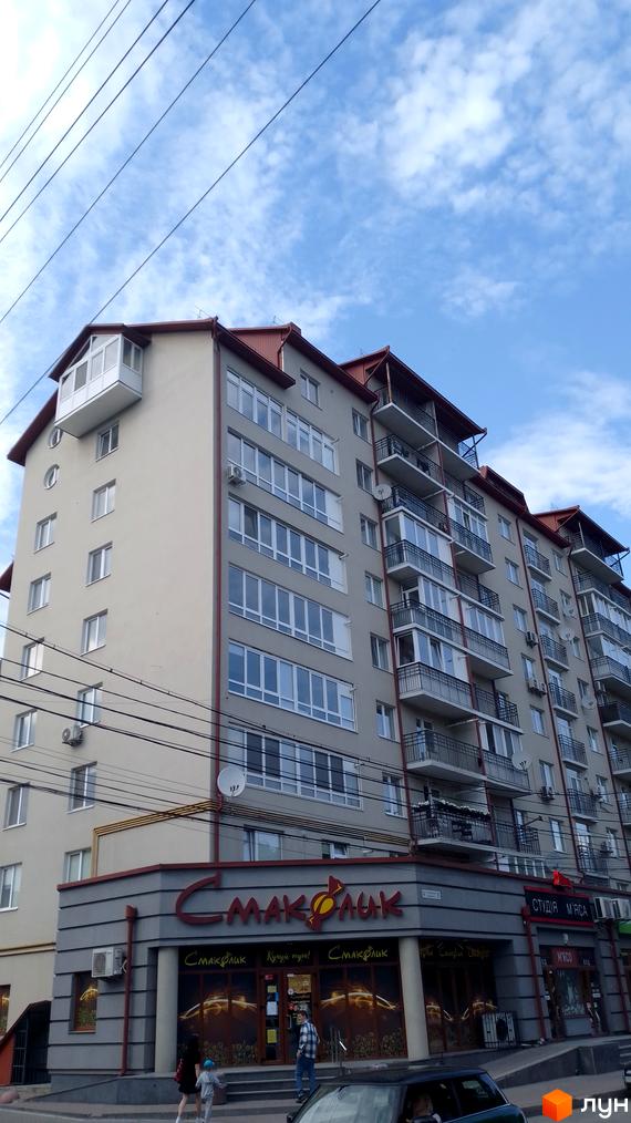 Зовнішній вигляд багатоповерхового будинку з червоним дахом та численними балконами. Фасад світло-сірого кольору, вікна великі, засклені.