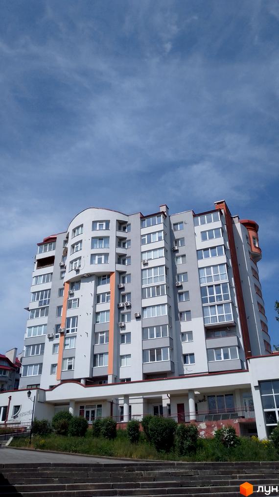 Зовнішній вигляд багатоповерхового будинку ЖК Співоче Поле з сірими та помаранчевими елементами фасаду. Навколо будинку зелені насадження та сходи.