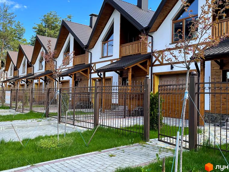 Зображення Котеджного містечка Desna Residence-2: ряд білих будинків з темними дахами, дерев'яними балконами та гаражами. На передньому плані видно металевий паркан, зелений газон та вимощені пішохідні зони.