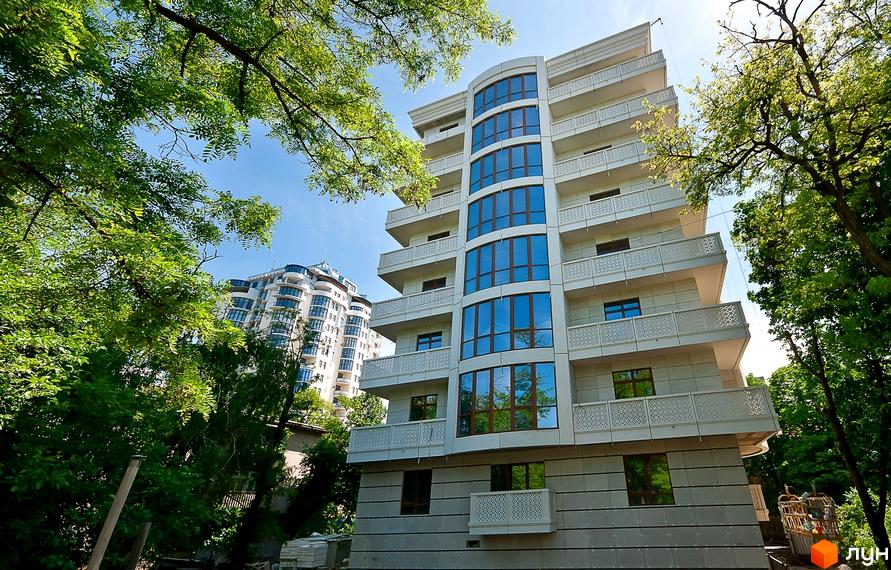 Зображення фасаду Житлового комплексу Біла акація з великими вікнами та балконами. Навколо будинку зелені дерева.