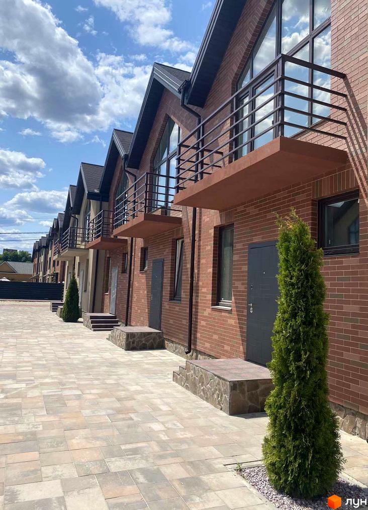 Цегляні таунхауси Modern House з балконами та темними дверима. Бруківка на території, зелені насадження та блакитне небо з хмарами.