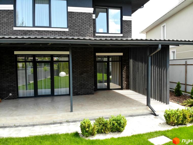 Фасад будинку Котеджного містечка Kulbaba Petropavlivka з темної цегли, великими вікнами та критою терасою. Прибудинкова територія має зелений газон та кущі.