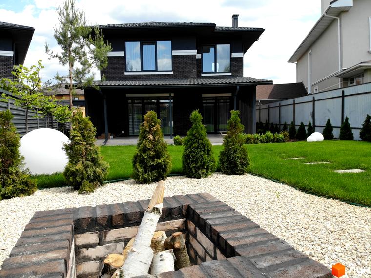 Темний двоповерховий будинок Котеджного містечка Kulbaba Petropavlivka з великими вікнами та доглянутим газоном, декоративними деревами і зоною для вогнища.