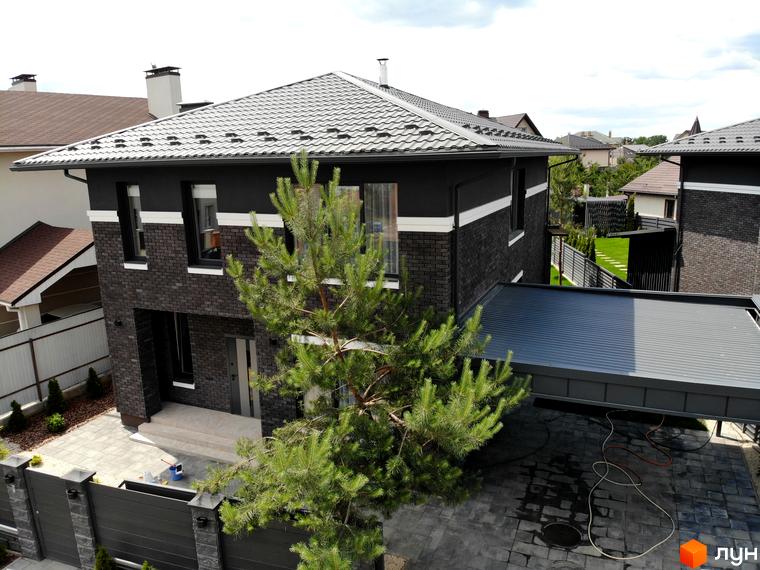 Котеджне містечко Kulbaba Petropavlivka: двоповерховий будинок з темної цегли та штукатурки, з сірою металочерепицею на даху. На подвір'ї бруківка, зелена сосна та навіс для авто.