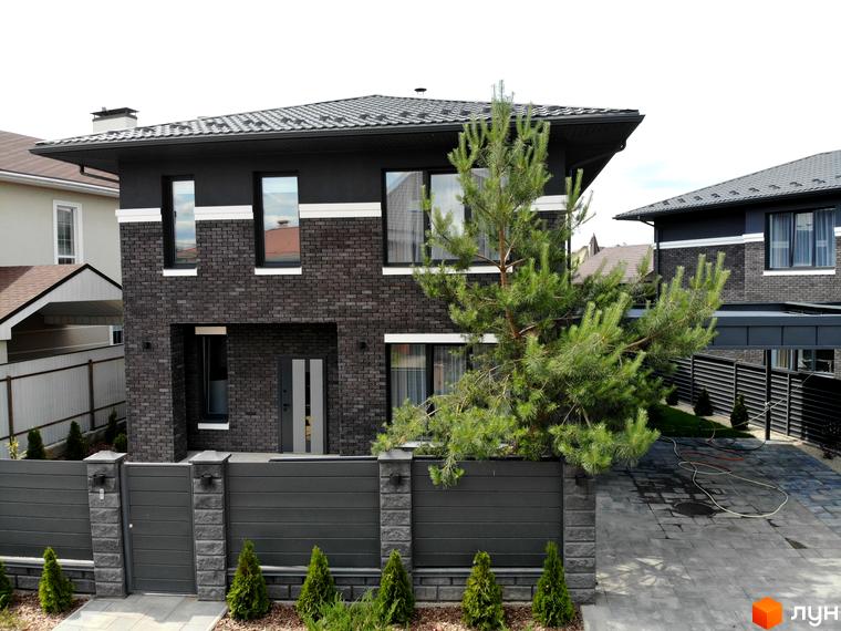 Двоповерховий будинок з темної цегли, білими смугами та великими вікнами. Прибудинкова територія Котеджного містечка Kulbaba Petropavlivka: сірий паркан, зелені насадження та брукований під'їзд.