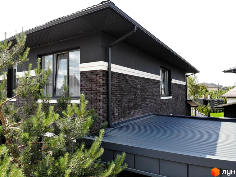 Фасад будинку Котеджного містечка Kulbaba Petropavlivka з темно-сірою цеглою та штукатуркою, білою смугою та темними вікнами. На передньому плані зелені хвойні дерева.