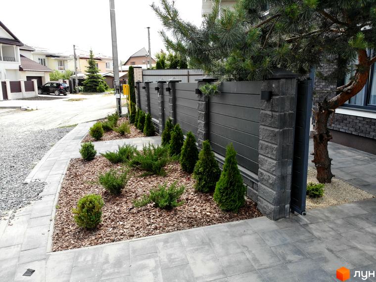 Котеджне містечко Kulbaba Petropavlivka: огорожа з темних горизонтальних панелей та кам'яних стовпів. Прибудинкова територія з доглянутими хвойними рослинами та бруківкою.