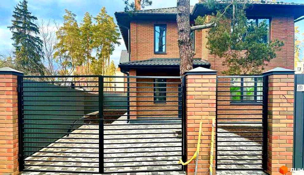 Двоповерховий цегляний будинок Котеджного містечка Villa Lisova з темним дахом та вікнами. Перед будинком чорна металева огорожа з цегляними стовпами та бруківка.