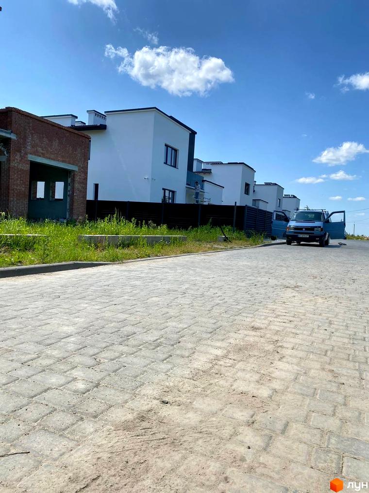 Будівництво котеджів у Котеджному містечку PRIDE Residents. Білі будинки з темними дахами та парканом, бруківка на дорозі, зелень.