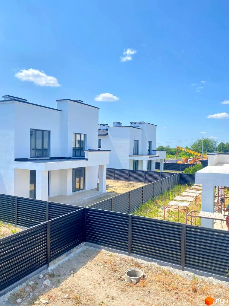 Білі будинки з темними вікнами та плоскими дахами, огороджені чорним парканом. На території видно суху траву та бетонні кільця.