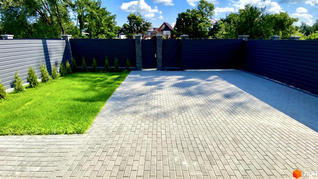 КМ Набережний: екстер'єр будинку з сірим парканом та воротами, облаштована прибудинкова територія з бруківкою та газоном.